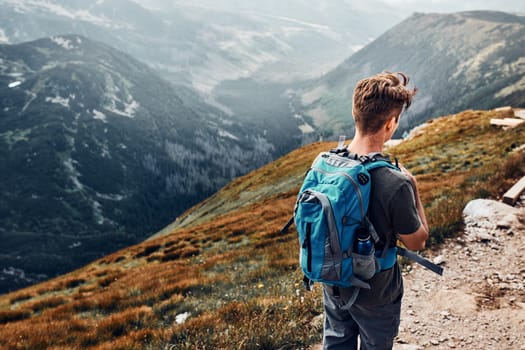 Young man with backpack hiking in a mountains, actively spending summer vacation. Rear view of teenager walking down from top of a hill along mountain path