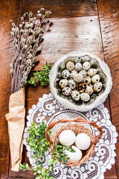 Easter composition of willow twigs with catkins, boxwood, quail eggs in a metal dish and easter basket with eggs, set on old wooden table. Catkins are wrapped up in paper. There is lacy napkin under the basket. Inside top shot. Vertical image.