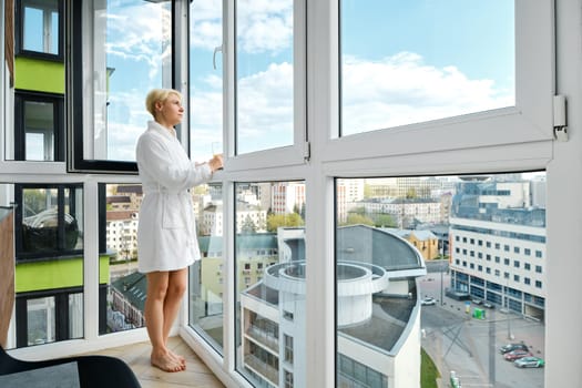 A mature, carefree woman stands barefoot in a bathrobe on her balcony enjoying an orange juice. Alone in her new high-rise apartment and overlooking the sunny city skyline.