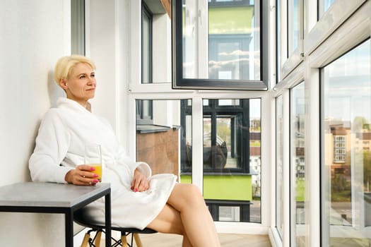 Middle aged woman in white bathrobe sitting on balcony of her new apartment with a glass of orange juice