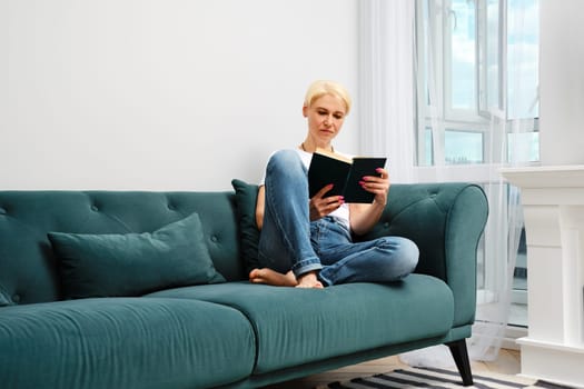 A middle aged woman with blond hair is relaxing on a sofa in her living room, reading a novel. She is dressed casually and enjoying the comfort of her home.