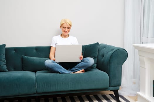 A mature woman relaxes on sofa while watching movies and browsing the web on her laptop, enjoying the comfort of independent domestic life.