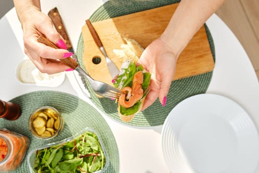 Top view of hands of middle aged woman puts salty salmon on sandwich while making breakfast