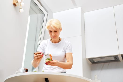 Low angle view of middle aged woman putting salty salmon on sandwich while making breakfast