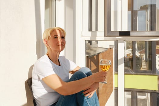 Middle-aged woman sits on balcony with glass of apple juice and squints in bright sun