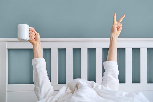 Man hiding under blanket showing hand with victory sign and holding cup of coffee. The early bird catches the worm concept.