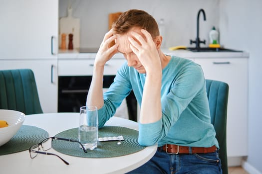 Sick middle aged man in the kitchen massaging head, the pills are on the table