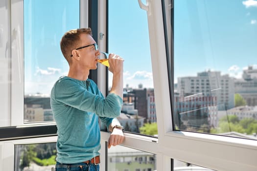 Middle aged man stands on balcony of high-rise building leaning against window frame and drinking juice