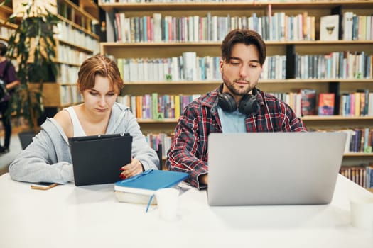 Students learning in university library. Young man preparing for test on laptop. Girl writing essay and making notes using computer. Focused students studying for college exams. Students discussing and learning together sitting at desk. Back to school