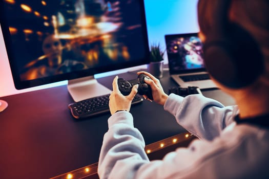 Teenager girl playing video game at home. Gamer holding gamepads sitting at front of screen. Streamer playing online in dark room lit by neon lights. Competition and having fun