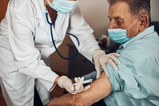 Doctor holding syringe with vaccine and making injection to senior patient with medical mask. Covid-19 or coronavirus vaccine. Physician wearing white coat and gloves using face mask