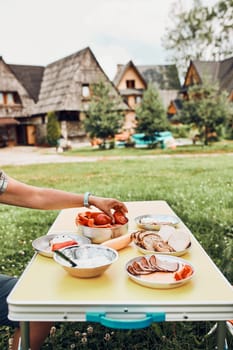Breakfast prepared during summer vacation on camping. Bread, cottage cheese, cold meat, tomatoes, fruits and coffee cups on table. Close up of outdoor table setting set on grass