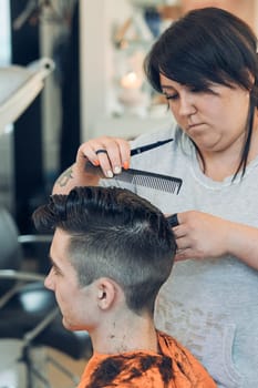 Hairdresser haircutting styling young man's hair in barbershop. Young woman working as a hairdresser in hair salon. Real people, authentic situations