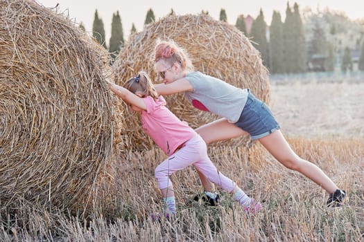 Sisters, teenage girl and her younger sister pushing hay bale playing together outdoors in the countryside. Candid people, real moments, authentic situations