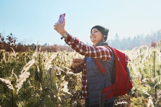 Young woman making video call sending greetings from vacation trip in mountains. Woman with backpack hiking through tall grass along path on meadow. Spending summer vacation close to nature