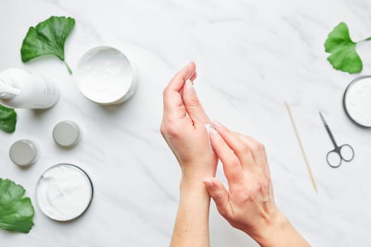 Woman applying  cosmetic moisturizing hand cream. Cosmetic products, green leaves on white table. Spa, manicure, skin care concept. Flat lay, overhead view