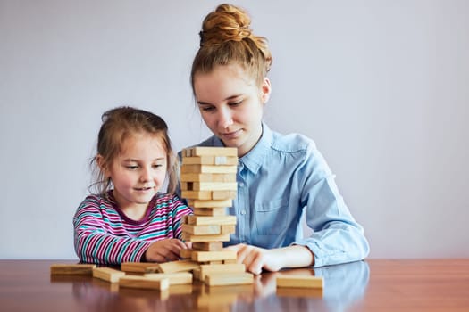 Little girl preschooler and her elder sister playing together with wooden blocks game toy. Siblings spending time sitting at desk at home