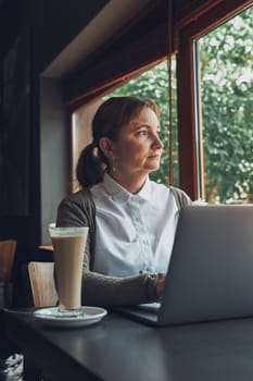Relaxed businesswoman working remotely on her laptop computer contemplate managing her work sitting at the table in a cafe
