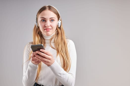 Young woman girl listening to music streaming content having fun watching video enjoying video chat talking with friends making gestures faces using smartphone earphones headphones standing over plain grey background