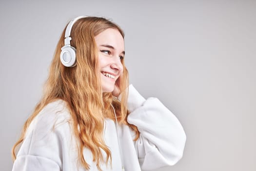 Young woman girl listening to music streaming content having fun watching video enjoying video chat talking with friends making gestures faces using smartphone earphones headphones standing over plain grey background