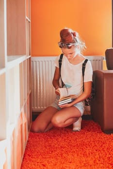 Teenage girl looking for books in local library. Young woman with a backpack sitting by a bookshelf indoors. Real people, authentic situations