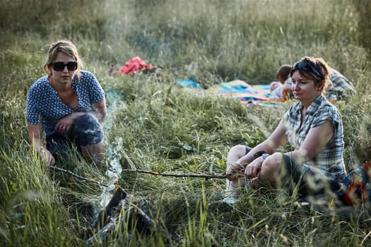 Women roasting a marshmallows over a campfire on meadow. Families spending vacations close to nature. Candid people, real moments, authentic situations