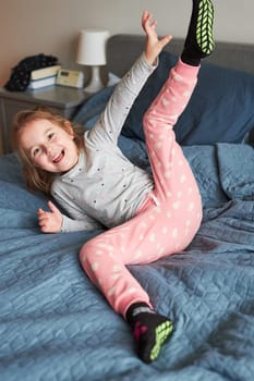 Little happy playful kid girl having fun making silly funny faces jumping on bed in bedroom in the morning at home