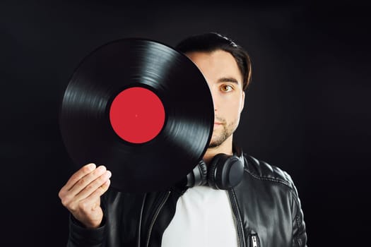 Man holding retro vinyl record covering face. Rock style. Vintage music style. Male wearing black jacket holding vinyl record disc standing on dark background. Retro music. Classic audio