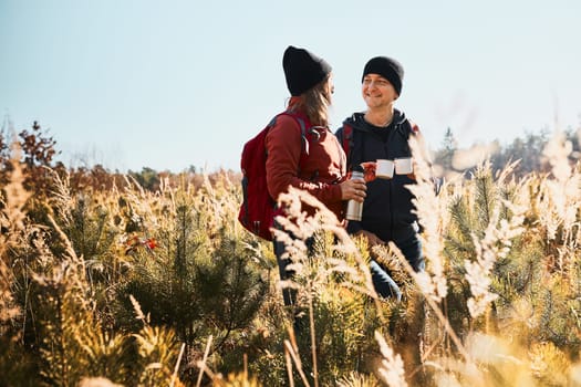 Friends relaxing and enjoying the coffee during vacation trip. People standing on trail drinking coffee. Couple with backpacks hiking through tall grass along path in mountains. Active leisure time close to nature