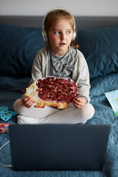 Little girl preschooler learning online showing her works drawings done at home. Child learning watching lesson remotely, talking with tutoress on video call from home during quarantine. Kid using laptop, headphones sitting on bed