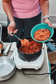 Close up of female hands putting rice dish with tomato sauce to a bowl. Woman cooking meal on electric stove on camping during summer vacations. Camping life, preparing a meal