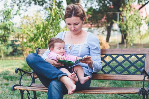 Mom reading a book and showing the pictures her little daughter sitting on bench in the garden