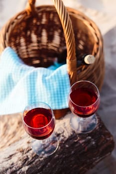 Two wine glasses with red wine standing on tree trunk, on beach, beside wicker basket with bottle of wine. View from above