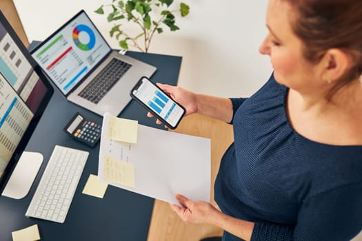 Businesswoman working with financial data on charts and tables on smartphone and computer. Woman entrepreneur looking at sale stats standing by desk in office