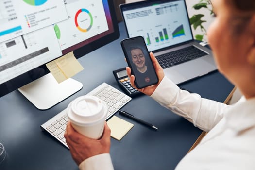 Woman entrepreneur having business video chat on smartphone. Businesswoman working with data on charts, graphs and diagrams on computer screen. Woman holding smartphone sitting at desk in office
