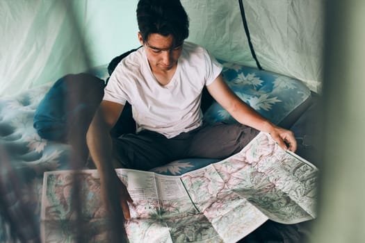 Young man looking at a map of mountain trails planning next trip on summer vacation sitting in a tent on camping. Concept of camp life