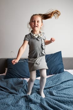 Little girl singing holding headphones cord imitating herself a real singer. Child having fun jumping dancing listening to music on bed in bedroom at home