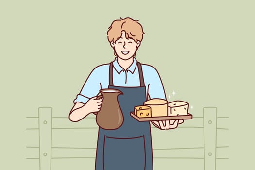 Man holds jug of milk and tray of cheese from cow farm selling own organic food. Guy wearing apron working on cow farm offers to buy organic dairy products with useful nutritional vitamins.