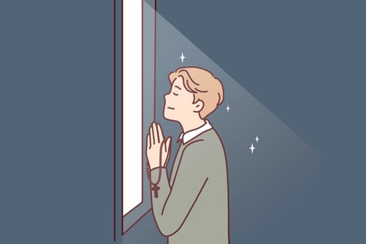 Religious man prays standing at window that emits light and holds chain with christian crucifix in hands. Believing guy prays turning to god is adherent of orthodox or catholic faith