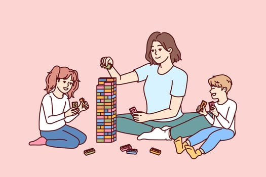 Woman babysitter together with kids plays constructor building tower of children blocks in kindergarten. Little boy and girl have good time and play educational games under supervision of babysitter