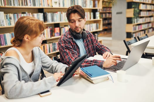 Students learning in university library. Young man preparing for test on laptop. Girl writing essay and making notes using computer. Focused students studying for college exams. Students discussing and learning together sitting at desk. Back to school