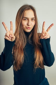 Excited girl gesturing peace sign. Portrait in positive mood. Girl making victory sign gesture. Beautiful model posing in studio over plain background looking at camera