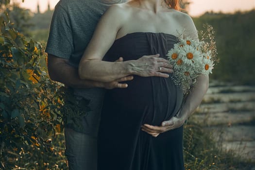 Pregnant woman and her husband hugging her tummy standing outdoors surrounded by nature. Pregnancy, expectation, motherhood concept