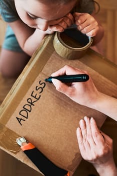 Woman writing an address on a cardboard box parcel in room at home. Little girl helping her mother to wrap a package. Preparing parcel to send