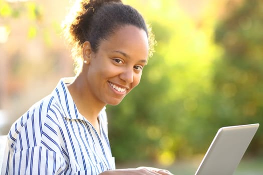 Happy black woman using laptop looking at you sitting in a park