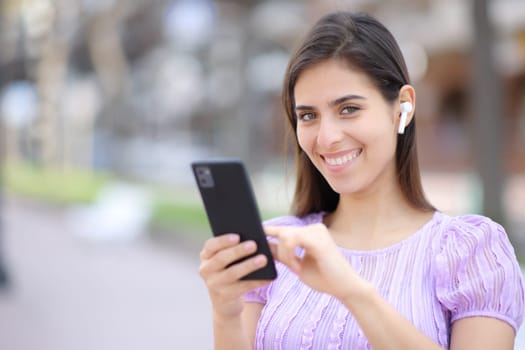 Happy woman posing looking at camera with earbud and phone in the street