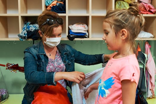 Child little girl changing clothes in changing room in nursery school with mothers help. Woman wearing face mask during pandemic coronavirus outbreak
