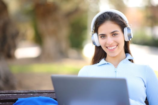 Happy student with laptop and headphone looks at you in a park