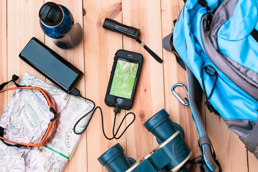 Charging a mobile phone during the trip in a mountains. Map, sunglasses, binoculars, backpack and other outdoor accessories on wooden table