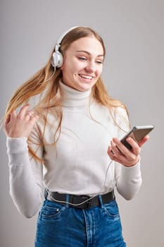 Young woman girl listening to music streaming content having fun watching video enjoying video chat talking with friends making gestures faces using smartphone earphones headphones standing over plain grey background